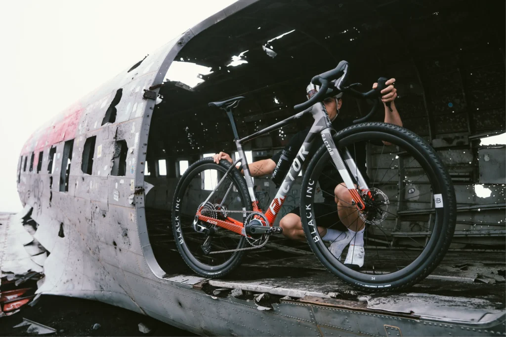A bicycle is being held by an obscured person, in a large opening of a broken airplane. The bike says the word 'factor' and the tires say 'black inc.'