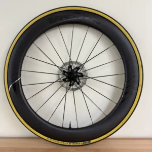 A bicycle wheel with yellow band of color, black spokes, leaning against a white wall on a wood floor.
